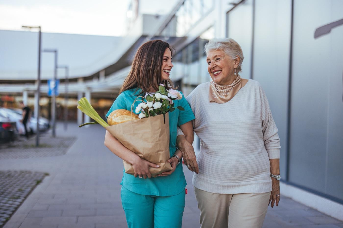 A caregiver walks alongside an older woman carrying groceries.