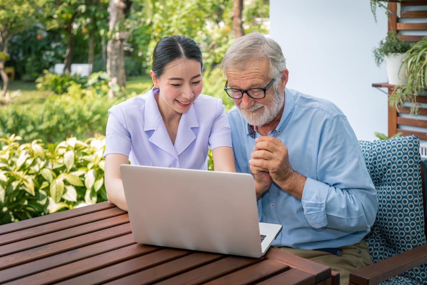 A caregiver and an older man sit together looking at a laptop.